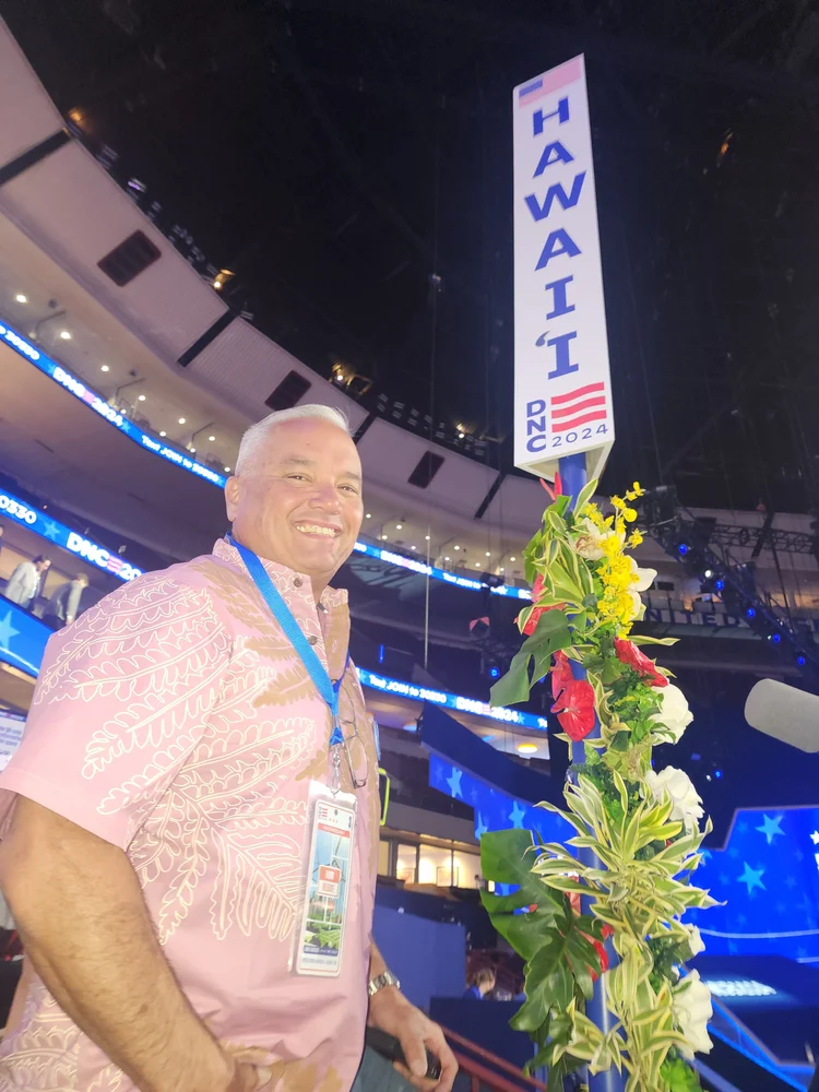 A man smiling at a political event inside a convention center with an American-themed sign that says 'Hawaii DNC 2024' and flowers in the foreground.