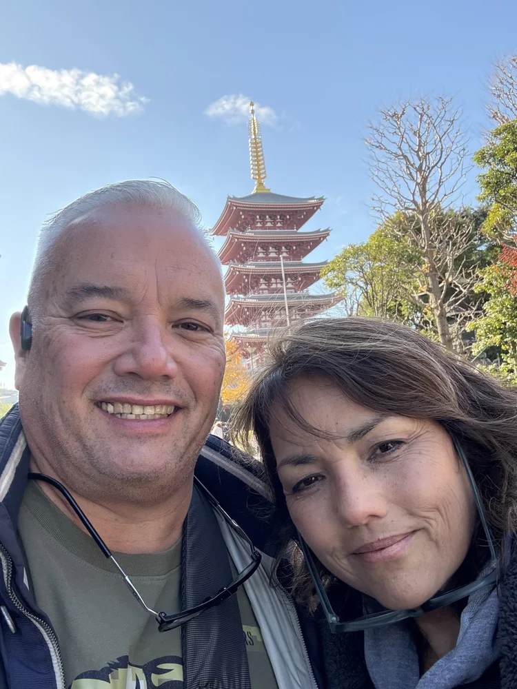 A smiling man and woman taking a selfie in front of a Japanese pagoda surrounded by trees, with a clear blue sky overhead.